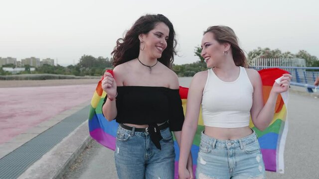 two young girls walking around holding hands and laughing with the LGBT flag of gay pride at sunset