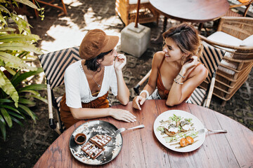 View from above at tanned girls in stylish outfits talking and enjoying tasty food in street cafe. Pretty woman in cap and lady in brown bra smile and eat waffle outside