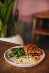 Vertical photo of baked croissant, tasty scrambled eggs, greens on pink plate on wooden table on blurred background