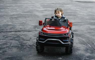 curly-haired boy in a striped T-shirt rides a red big toy car driving on an asphalt path. day off, outdoor recreation