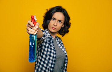 Stay where u are. Close-up photo of a strict pretty girl, in a checkered shirt, pointing a blue bottle spray on a camera , looking with a threat.