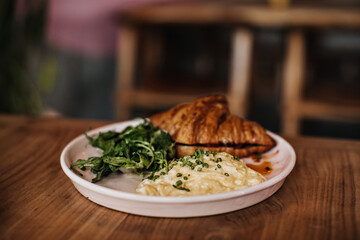 Photo of tasty baked croissant, scrambled eggs, greens on pink plate on wooden table table on blurred background