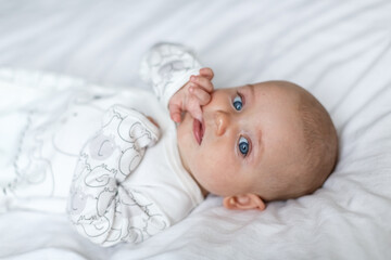 A little baby is smiling lying on the bed on a white sheet in the parents bedroom. A white jumpsuit in newborn. Cute baby lying on the bed. Closeup portrait of newborn baby looking at camera