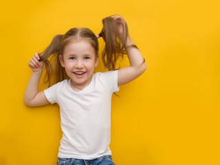 A naughty little girl in a white T-shirt is holding herself by the hair on a yellow background....