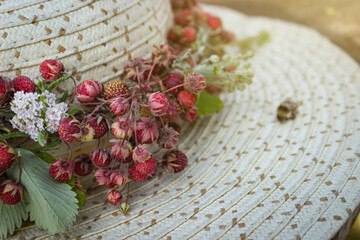 Bunches of wild berries on a light straw hat. Fresh, ripe wild strawberry, flowers and leaves on summer hat. Close up, copy space, postcard. Summer outdoor concept.
