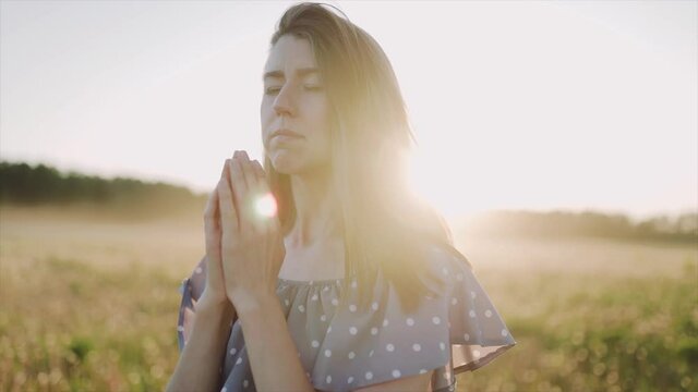 A caucasian beautiful wonan standing on the beautiful field during summer sunset and pray and look at the sky. The woman wears beautiful dress. Close up. SLOW MOTION