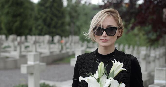 Close up of woman in dark glasses raising head and looking to camera. Young widow holding white lily flower while standing at cemetery. Concept of memorial day. Blurred background