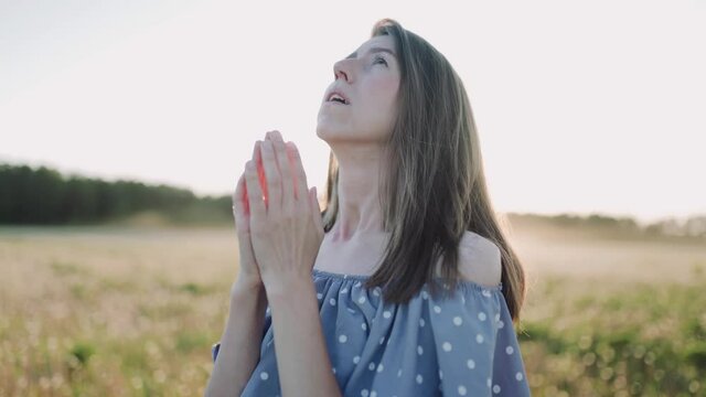 A caucasian beautiful wonan standing on the beautiful field during summer sunset and pray and look at the sky. The woman wears beautiful dress. Close up.