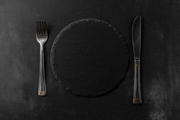 Round empty black granite stone board with shiny fork and knife top view. black textured cement background and template with copy space for your text