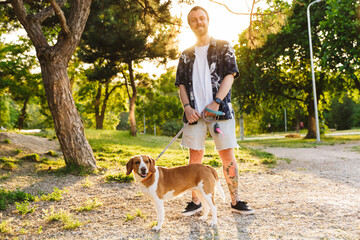 Image of young man smiling while walking with beagle dog in summer park