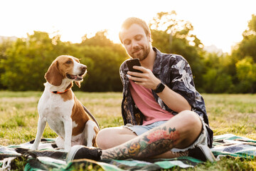 Image of young man using cellphone and sitting with beagle dog in park