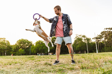 Fototapeta premium Image of young man smiling and playing with his beagle dog in park