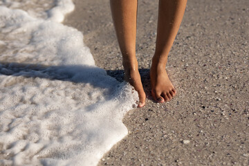 A mixed race woman on beach on a sunny day with a wave touching her bare feet 