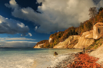 Ruegen Island with the chalk cliffs in the morning. Coastline in autumn at sunrise with sunshine. Trees and stony coast with foliage and clouds in waves of the Baltic Sea