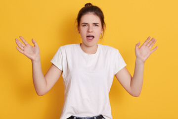 Angry emotional female with dark hair and knot, spreading hands aside and screaming, standing against yellow wall, dresses white casual t shirt, keeps mouth opened.