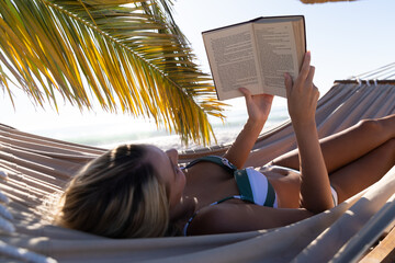 Caucasian woman lying on a hammock and reading a book at the beach.