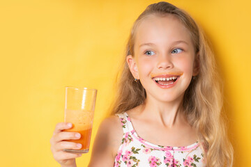 Smiling little girl in summer clothes looking to the side, holding a glass of fresh carrot juice in her hand. Waist shot isolated on yellow, copy space. mustache from carrot juice