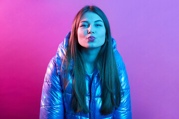 Young woman with long hair keeping lips rounded, making kiss,looking directly at camera, wearing jacket, posing isolated over pink neon background, woman expresses positive.