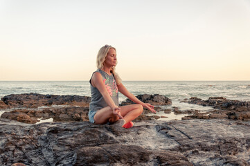 Fototapeta na wymiar A blonde girl in a T-shirt and shorts sits with her eyes closed on the rocky shore of the sea and meditates during sunset.