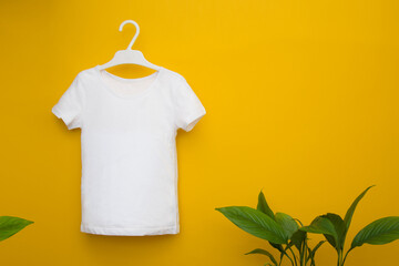 White children's t-shirt hanging on a yellow background next to green leaves. Mocap. Copyspace