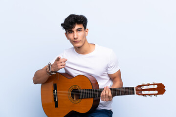 Young Argentinian man with guitar over isolated blue background surprised and pointing front