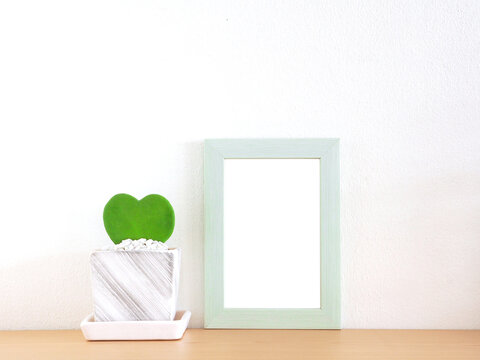 Green border photo frame and heart-shaped tree in a flower pot placed on a wooden shelf with a white wall as the background...