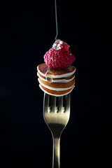 Tiny cereal pancakes with raspberries strung on a fork with pouring honey on a dark background
