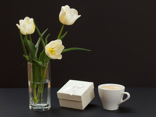 Bouquet of yellow tulips in vase with gift box and cup on a black background.