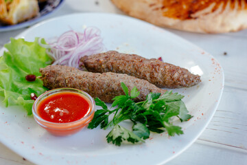 Meat kebabs on white plate with green herbs and red sauce.