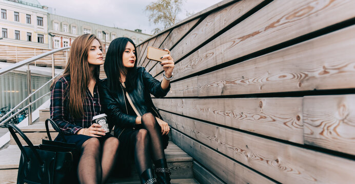 Two women making photo on cell telephone while sitting on a wooden stairs in the fresh air, young hipster girls photographing herself on mobile camera while relaxing after walking, copy space area