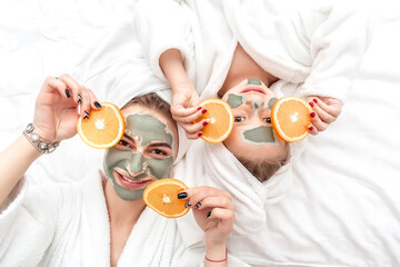 Happy mother and child daughter make face skin mask holding a slice of orange in front of theirs...
