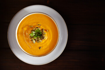Vegetarian autumn pumpkin soup puree with seeds, greenery and oil in white bowl on black background. Top view.