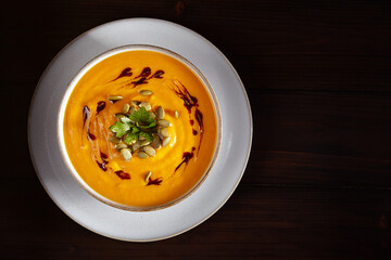 Vegetarian autumn pumpkin soup puree with seeds, greenery and oil in white bowl on black background. Top view.