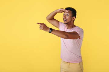 Joyful african american young man looking far away pointing finger over yellow background.