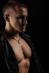 Handsome man wearing leather jacket on a naked muscular torso, on a dark background, looking straight.