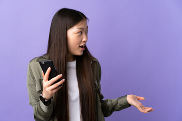 Young Chinese girl using mobile phone over isolated purple background with surprise facial expression