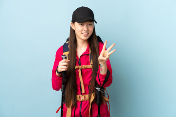Young Chinese girl with backpack and trekking poles over isolated blue background happy and counting three with fingers