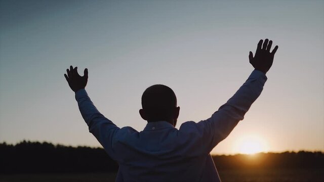A caucasian man standing on the beautiful field during summer sunset, his hands are raised. A man look at the sky and pray. A man stand with his back to the camera. Close up. SLOW MOTION