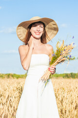 Young beautiful woman in a white dress and a hat holds a bouquet with wildflowers on a millet field. Concept of outdoor recreation, a trip to the village
