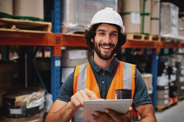 Handsome cheerful young caucasian smiling supervisor with white helmet using digital tablet for work while standing in warehouse against goods