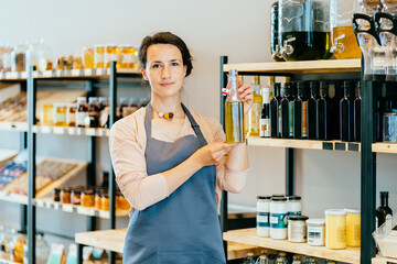 Young attractive woman working in a grocery store, female owner holding glass bottle with olive oil over shelves with eco packaging free products working in a zero-waste shop.