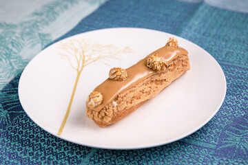 Eclair with caramel glaze lays on the white plate. Turquoise tablecloth under the plate.