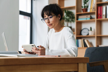Photo of focused woman using smartphone while working with laptop