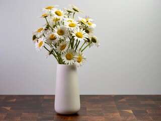 White vase full of fresh cut daisy white and yellow wildflowers sitting on a cutting board surface with a white wall behind.  Nature brought inside for still life photography.