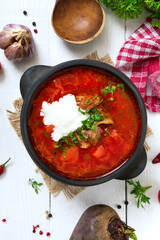 Traditional Ukrainian borsch with young vegetables and meat in a black clay pot on a white wooden background. Top view.