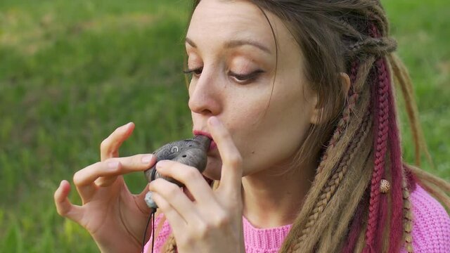 Portrait of young female shaman in pink knitted sweater playing on ceramic ocarina in the forest during autumn or spring. Relaxing tranquil scene, traditional music concepts