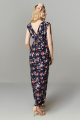 Turned back lady in high heel shoes is wearing dark blue summer jumpsuit with V-neckline and colorful flower print. The lady's blonde hair is decorated with a pink and white flower chaplet.