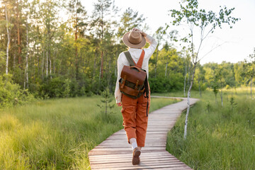 Woman botanist with backpack on ecological hiking trail in summer outdoors. Naturalist exploring wildlife and ecotourism adventure walking on path through peat bog swamp in a wildlife national park. 