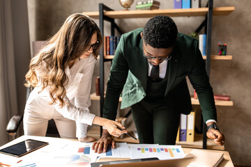 Work in a creative office. An African man and a beautiful business lady in formal clothes are working together, standing indoors