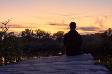 A man sitting alone on the old wooden bridge near the small countryside river and enjoying sunset and coffee in plastic glass on a calm evening